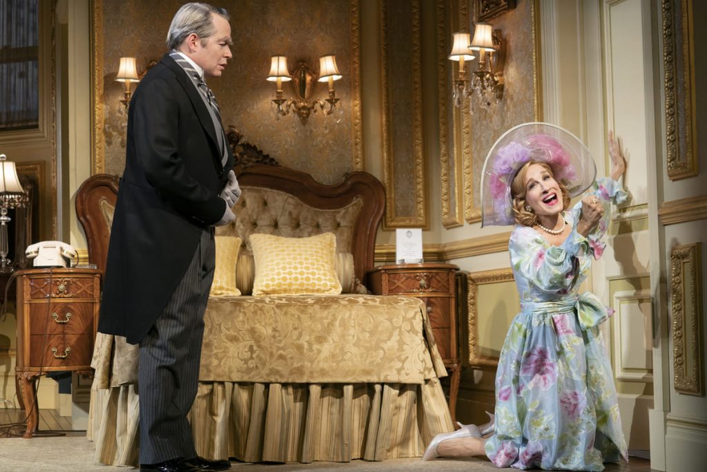 Sarah Jessica Parker kneels in front of and is going to bang her fist against a closed door. She wears a floral blue dress with a wedding facinator hat on. Matthew Broderick stands to the left in a 3 piece suit. Behind them is an ornate bed.