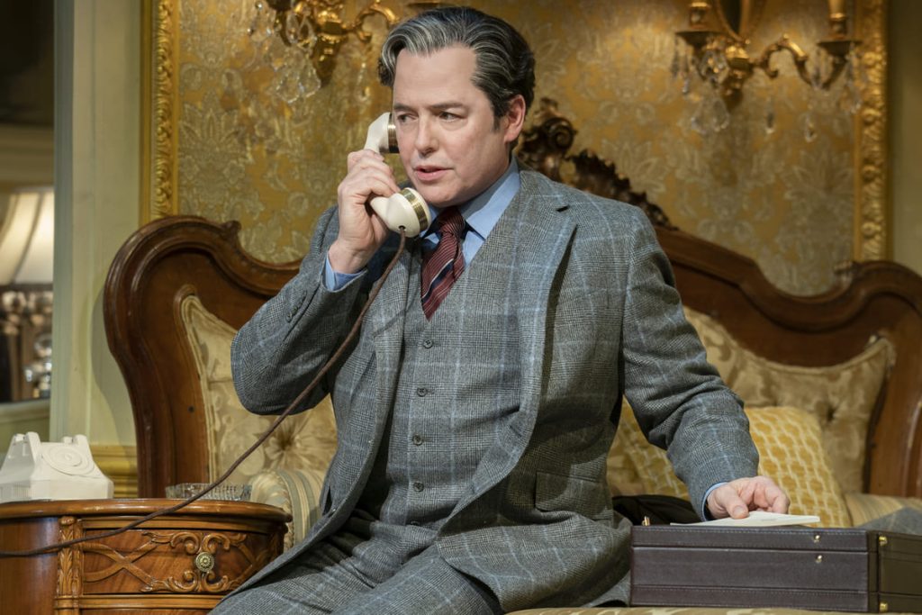 Matthew Broderick wears a workwear-esque suit. He sits on an ornate bed and is on the phone. Next to him is a briefcase.